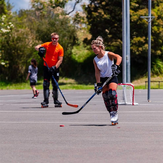 Two people wearing inline skates on an outdoor bitchumen court holding hockey sticks and moving a hockey puck with a red and white goal in the background